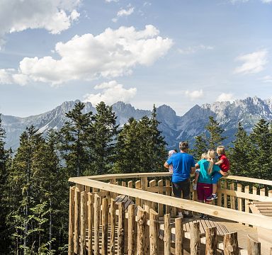 Aussichtsplattform mit Blick auf bewaldete Hügel und imposante Bergkette im Hintergrund, Menschen genießen die Landschaft an einem sonnigen Tag.