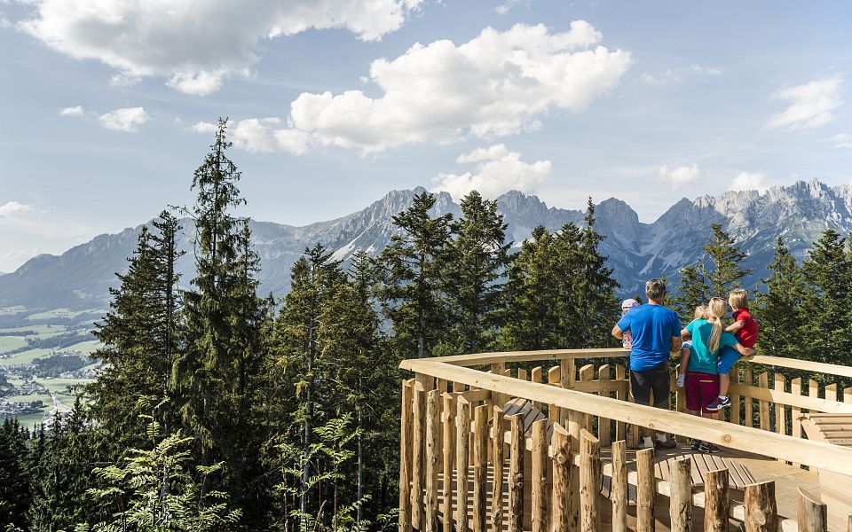 Aussichtsplattform mit Blick auf bewaldete Hügel und imposante Bergkette im Hintergrund, Menschen genießen die Landschaft an einem sonnigen Tag.
