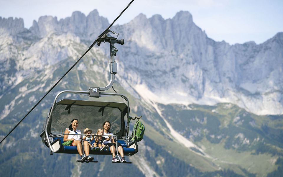 Familie sitzt in einem Sessellift vor einem markanten Bergmassiv, umgeben von grünen Wiesen und einem klaren blauen Himmel, im Alpengebiet Österreichs.