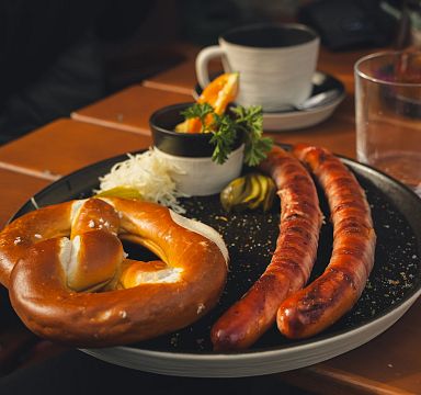 Eine Nahaufnahme eines Tellers mit zwei Bratwürsten, einer Brezel, Sauerkraut und Pickles. Im Hintergrund ist eine Tasse auf einem Holztisch zu sehen.