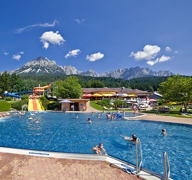 Outdoor pool with people swimming, surrounded by greenery and mountains in the background under a clear blue sky at the Wilder Kaiser region.