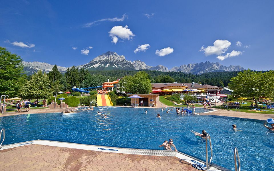Outdoor pool with people swimming, surrounded by greenery and mountains in the background under a clear blue sky at the Wilder Kaiser region.