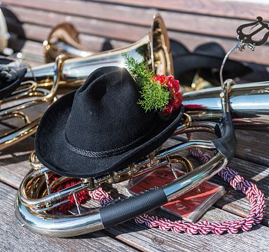 Auf einer Holzbank liegen Blasinstrumente, umgeben von schwarzen Tiroler Hüten mit roten Blumen und grünen Tannenzweigen als Dekoration.