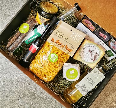 Ein Geschenkkorb mit regionalen Produkten: Nudeln, Wurst, Käse, Holunderblütensirup, Öl und Brot auf Heubett arrangiert, präsentiert eine kulinarische Vielfalt.