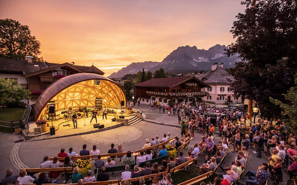 Outdoor concert at sunset near Wilder Kaiser, with mountains in the background and an illuminated stage. Audience seated on steps, surrounded by charming buildings.