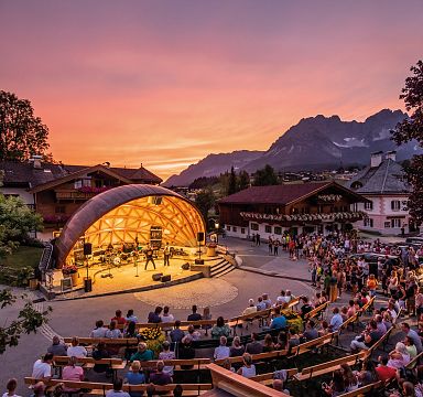 Ein Open-Air-Konzert in einem Bergdorf bei Sonnenuntergang. Zuschauer sitzen vor einer beleuchteten Bühne, umgeben von traditionellen Gebäuden und Bergen im Hintergrund.
