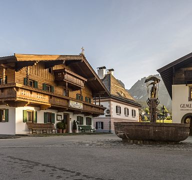 Ein malerischer Dorfplatz mit traditionellen Tiroler Holzhäusern und einem Brunnen in der Mitte, umgeben von Bergen und blauem Himmel im Hintergrund.