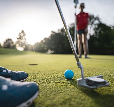 Nahaufnahme eines Golfschlägers und Balls auf dem Grün, mit unscharfer Person im Hintergrund, die einen Golfschläger hält. Sonnenschein im Hintergrund.