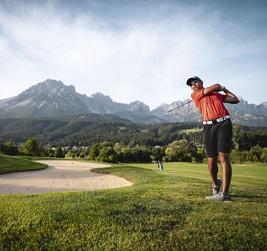 Ein Golfspieler in rotem Shirt schlägt einen Ball auf einem Rasenplatz mit sandigem Bunker, im Hintergrund majestätische Berge unter blauem Himmel.
