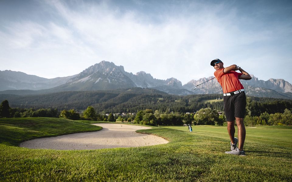 Ein Golfspieler in rotem Shirt schlägt einen Ball auf einem Rasenplatz mit sandigem Bunker, im Hintergrund majestätische Berge unter blauem Himmel.