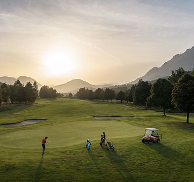 Golfers play at sunrise on a lush green course with distant Wilder Kaiser mountains, bathed in golden morning light. A golf cart is parked nearby, and trees line the fairway.