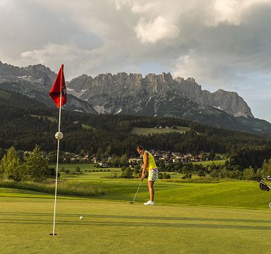 A golfer lines up a putt on a green with a picturesque mountain range in the background and a red flag near the hole.