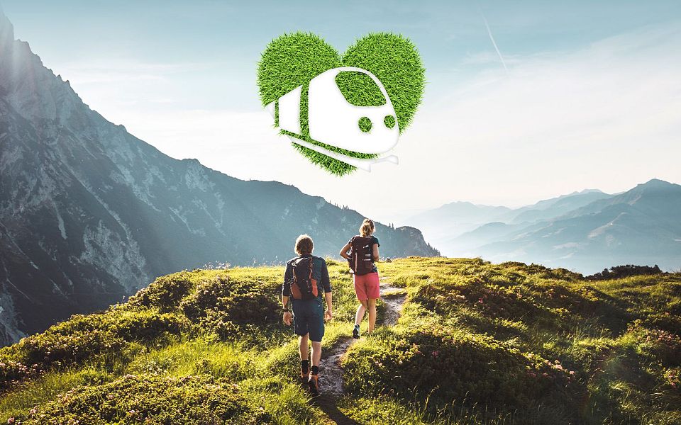 Two people hike on a green mountain trail with the Wilder Kaiser mountains in the background, under a blue sky with a heart-shaped eco symbol overhead.