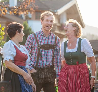 Three people in traditional attire laughing outdoors with a blurred background of trees and a house.