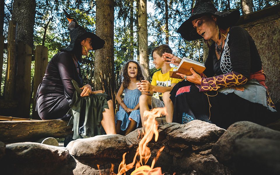 Zwei Erwachsene in Hexenkostümen erzählen Kindern am Lagerfeuer im Wald Geschichten. Die Szenerie wirkt verspielt und magisch in einer natürlichen Umgebung.