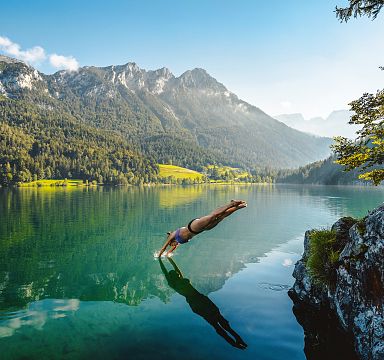Person taucht in klaren Bergsee mit Wald und Bergen im Hintergrund. Sonniges Wetter, ruhige Atmosphäre, grüne Umgebung und malerische Landschaft.
