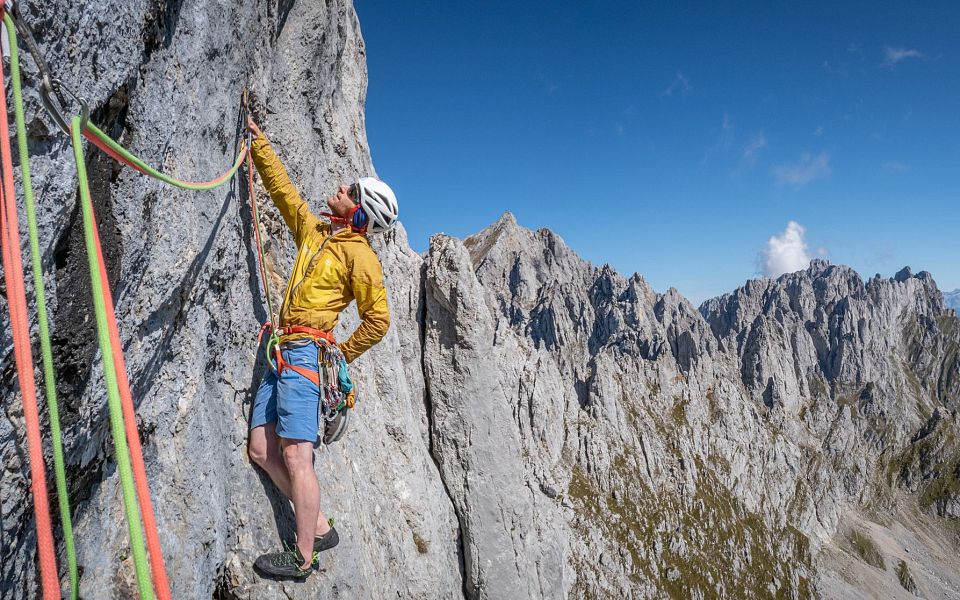 Kletterer in gelber Jacke am felsigen Bergmassiv des Wilden Kaisers, blauer Himmel im Hintergrund. Spektakuläre Berglandschaft in Tirol.