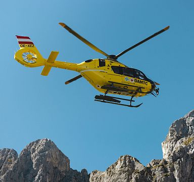 Gelber Rettungshubschrauber fliegt vor blauem Himmel über felsigem Bergmassiv in den Alpen. Österreichische Flagge am Heck des Helikopters sichtbar.