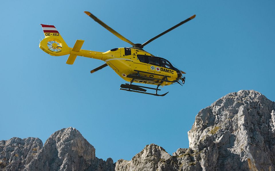 Gelber Rettungshubschrauber fliegt vor blauem Himmel über felsigem Bergmassiv in den Alpen. Österreichische Flagge am Heck des Helikopters sichtbar.