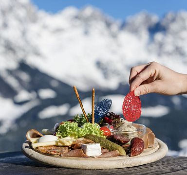 A platter of Tyrolean snacks including cheese and sausage is set on a table with the snow-covered peaks of the Wilder Kaiser mountains in the background.