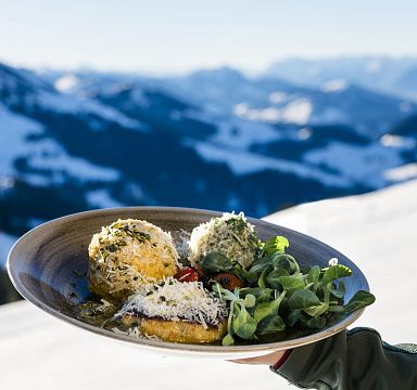 Plate of gourmet food with fresh greens, set against a backdrop of snow-capped Wilder Kaiser mountains, merging fine dining with scenic alpine views.