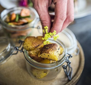 Nahaufnahme einer Hand, die ein dekoratives Blumenarrangement auf einem Gericht mit gebratenen Kartoffeln platziert. Im Hintergrund sind weitere Speisen zu sehen.