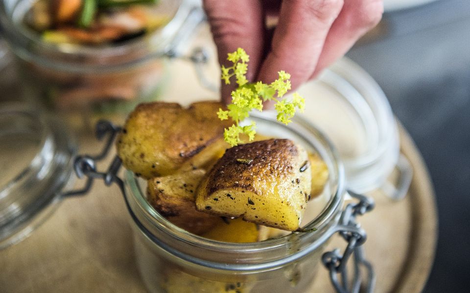 Nahaufnahme einer Hand, die ein dekoratives Blumenarrangement auf einem Gericht mit gebratenen Kartoffeln platziert. Im Hintergrund sind weitere Speisen zu sehen.