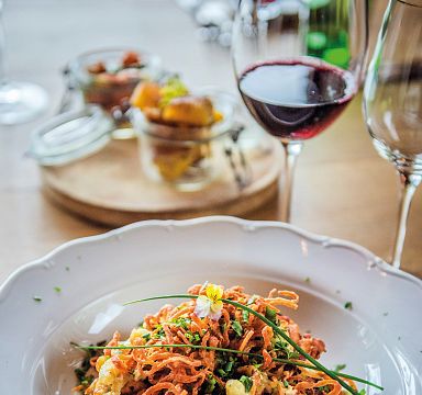 Ein Teller mit traditionellem Gericht, vermutlich Tiroler Gröstl, garniert mit Zwiebeln und Kräutern. Im Hintergrund ein Glas Wein auf einem Tisch.