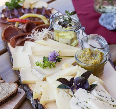 Ein rustikales Holzbrett mit verschiedenen Käsesorten, salamiähnlicher Wurst, zwei Gläsern mit Dip und garnieren Kräutern dekoriert.