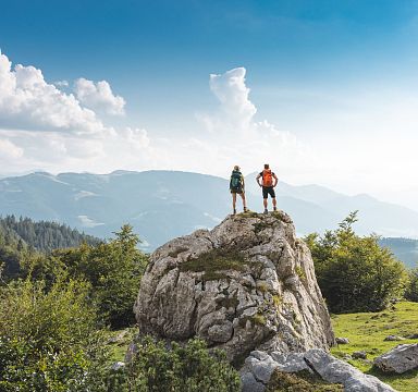 Zwei Wanderer stehen auf einem großen Felsen in einer bergigen Landschaft. Sie genießen die Aussicht auf grüne Hügel und blauen Himmel mit weißen Wolken.