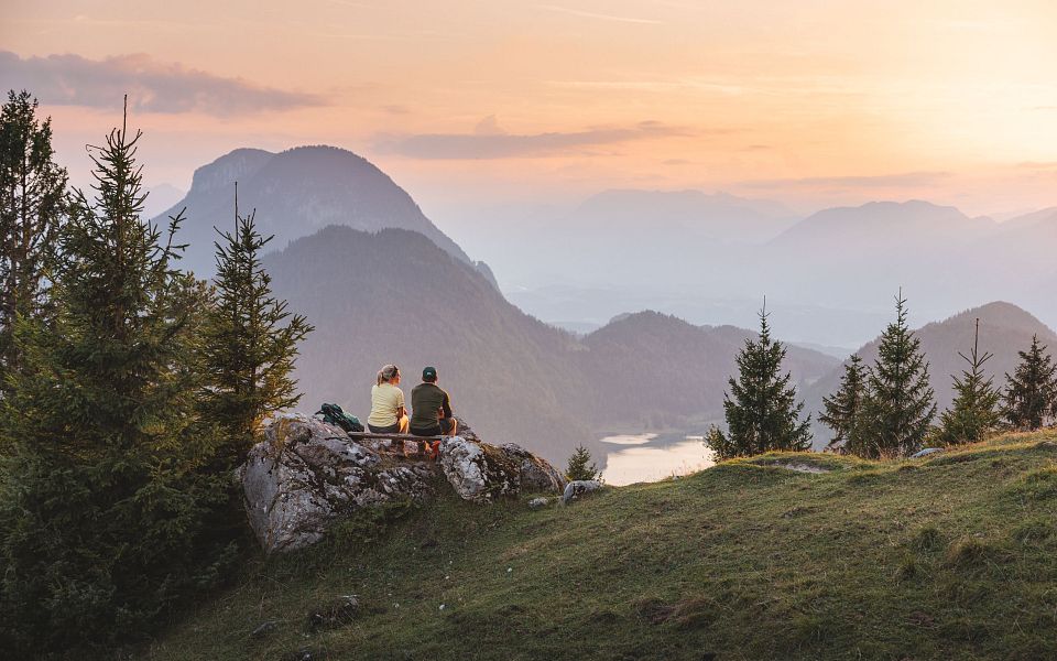 Zwei Personen sitzen auf einem Hügel, genießen den Sonnenuntergang mit Blick auf bewaldete Berge und einen klaren Himmel. Die Atmosphäre ist friedlich und idyllisch.