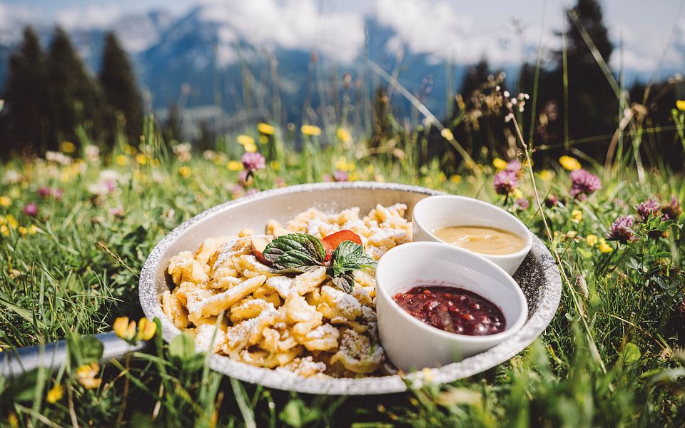 Ein Teller Kaiserschmarrn mit Puderzucker, Apfelmus und roter Marmelade steht auf einer Wiese mit Blumen im Vordergrund und Bergen im Hintergrund.