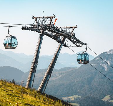 Moderne Gondelbahn fährt über grasbewachsene Hügel, mit beeindruckendem Bergpanorama im Hintergrund unter klarem, blauem Himmel.