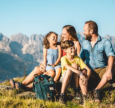 Eine vierköpfige Familie sitzt lachend auf einer Wiese vor einem imposanten Bergpanorama. Der Himmel ist klar und die Sonne scheint.
