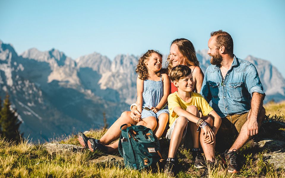 Eine vierköpfige Familie sitzt lachend auf einer Wiese vor einem imposanten Bergpanorama. Der Himmel ist klar und die Sonne scheint.