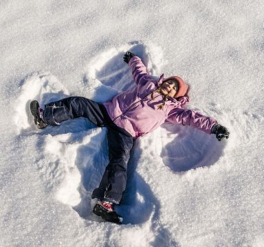 Ein Kind in rosa Jacke und schwarzer Hose liegt im Schnee und macht einen Schneeengel. Der Himmel ist nicht sichtbar, nur der weiße, glitzernde Schnee.