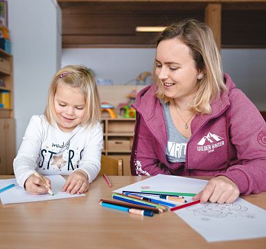 Eine Frau und ein kleines Mädchen zeichnen zusammen an einem Tisch. Sie sind entspannt und lächeln, umgeben von bunten Buntstiften und Zeichnungen.