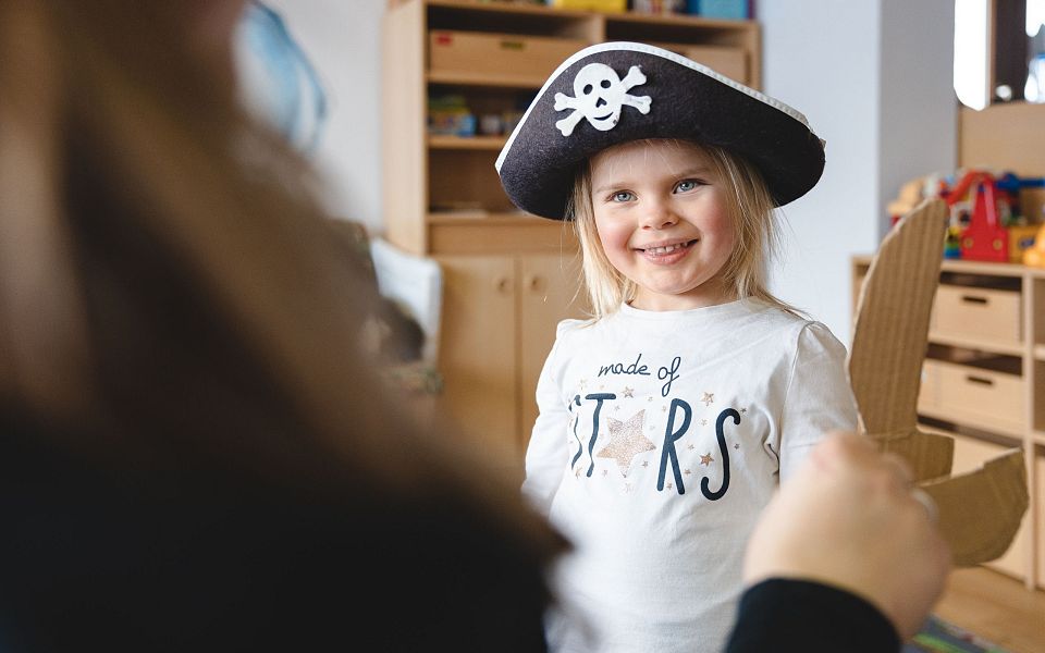 A young child in a pirate hat smiles in a playroom filled with toys and shelves, engaging in imaginative play with an adult nearby.