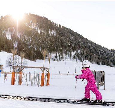 Ein Kind in pinkem Skianzug fährt auf einem Förderband im Schnee, umgeben von verschneiten Hügeln und Wald, während die Sonne hinter einem Berg aufgeht.