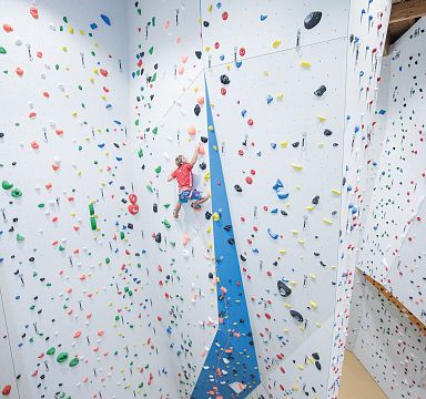 Kind klettert in einer Kletterhalle an einer bunt markierten Wand mit zahlreichen Griffen und Tritten, umgeben von hellen Wänden und blauen Akzenten.