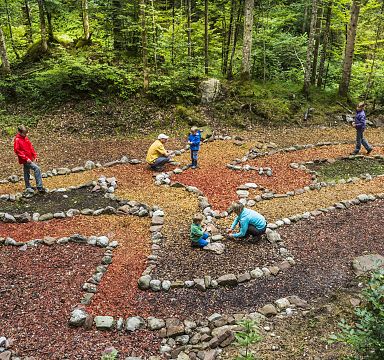 Menschen laufen in einem Steinlabyrinth im Wald, umgeben von Bäumen. Die Wege sind mit verschiedenen farbigen Blättern ausgelegt. Es herrscht eine entspannte Atmosphäre.