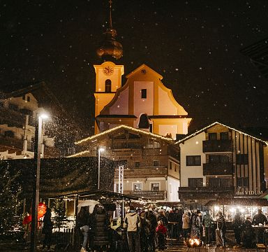 A lively winter market scene at Wilder Kaiser. People gather under softly falling snow near warmly lit traditional buildings and a church, enjoying the festive atmosphere.
