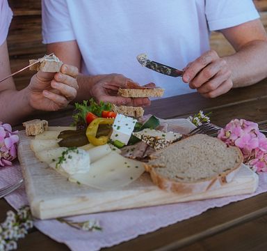 Zwei Personen genießen eine Jause mit Käse, Brot und Wein. Eine Holzplatte mit verschiedenen Käsesorten und Gemüse liegt auf einem Holztisch, umgeben von Blumen.