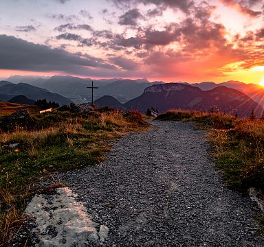 Sonnenuntergang in den Bergen mit einem Bergkreuz und einem schmalen Wanderweg im Vordergrund. Dramatische Wolken und warme Farben am Himmel.