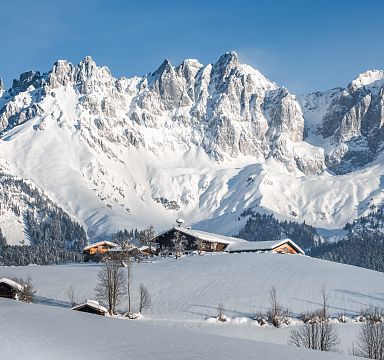 Schneebedeckte Alpenlandschaft mit beeindruckendem Bergmassiv im Hintergrund, Holzhütten im Vordergrund, blauer Himmel, klare Winteratmosphäre.