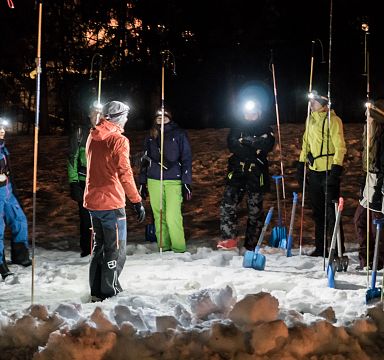 Menschen stehen bei Nacht im Schnee in einer Gruppe, ausgestattet mit Stirnlampen und Schneeschaufeln, offenbar in einer Art Training oder Übung.