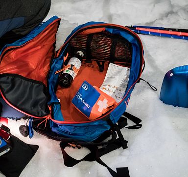 Ein geöffneter Rucksack liegt auf Schnee, gefüllt mit einer Flasche, einem Notizbuch und Ausrüstung. Daneben eine Schaufel und ein zusammenklappbarer Stock.