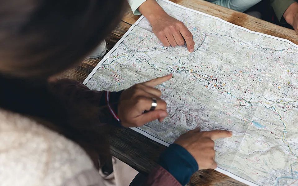 Personen schauen auf eine Landkarte auf einem Tisch, während sie mit Fingern Richtungen zeigen. Eine entspannte Outdoor-Szene in einem natürlichen Setting.
