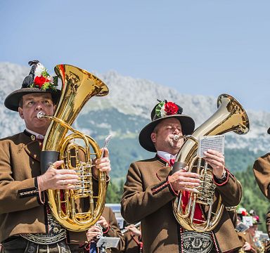 Blaskapelle in traditioneller Tiroler Tracht spielt Musikinstrumente vor Bergkulisse in der Region Wilder Kaiser an einem sonnigen Tag.