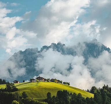 View of the Wilder Kaiser mountains partially covered in clouds with green hills and a farmhouse in the foreground in Tirol.